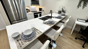 Una cocina moderna con un refrigerador de acero inoxidable, un microondas y una larga mesa de comedor dispuesta con platos, cubiertos y vasos blancos y azules.