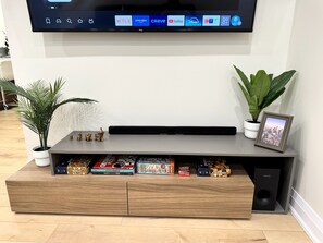 Ein Flachbildfernseher an der Wand, eine Holz-Unterhaltungsecke mit Büchern und Spielen, zwei Blumentöpfe und ein gerahmtes Bild.
