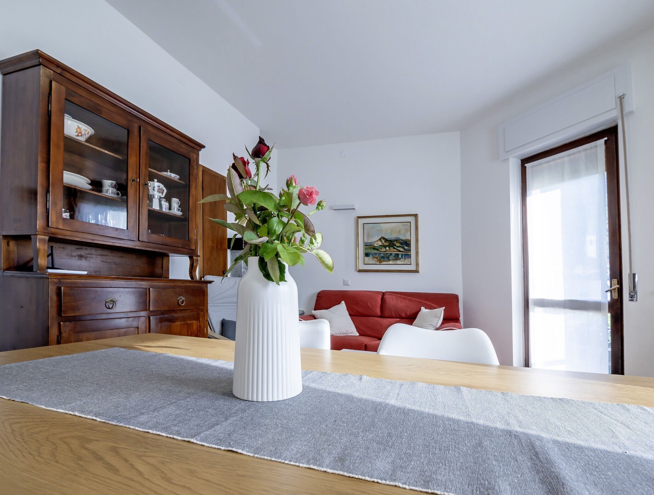 A room with a wooden table, a white vase with flowers, a red sofa, and a cabinet with glass doors.