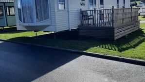 Ein Mobilheim mit Veranda und Deck, gelegen auf einer grünen Wiese unter blauem Himmel.