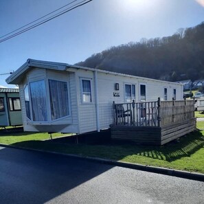 Ein Mobilheim mit Veranda und Deck, gelegen auf einer grünen Wiese unter blauem Himmel.