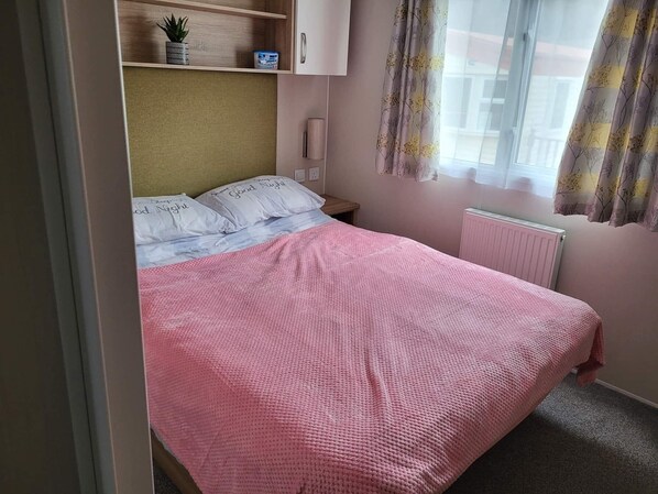 Ein Schlafzimmer mit Bett, Kissen, einem Nachttisch, einem Regal mit Pflanze, einer Heizung und einem Fenster mit Vorhängen.