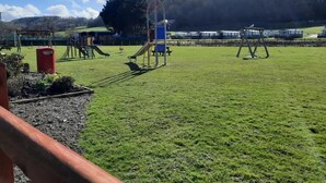 Ein Spielplatz mit Schaukeln und Rutschen, einem roten Mülleimer und einer grünen Wiese.