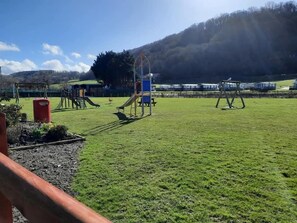 Ein Spielplatz mit Schaukeln und Rutschen, einem roten Mülleimer und einer grünen Wiese.