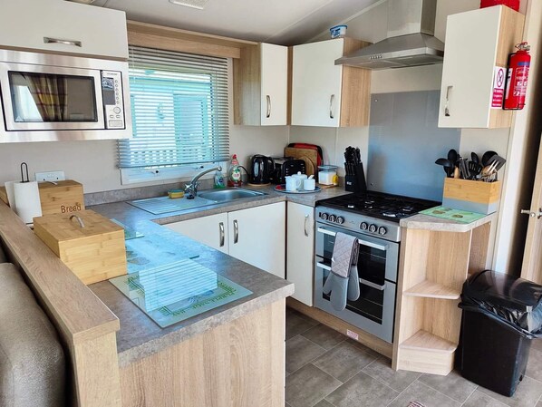 A compact kitchen with a microwave, stove, oven, sink, and wooden cabinets. - Clarach Bay Holiday Village W18 (Aberystwyth)