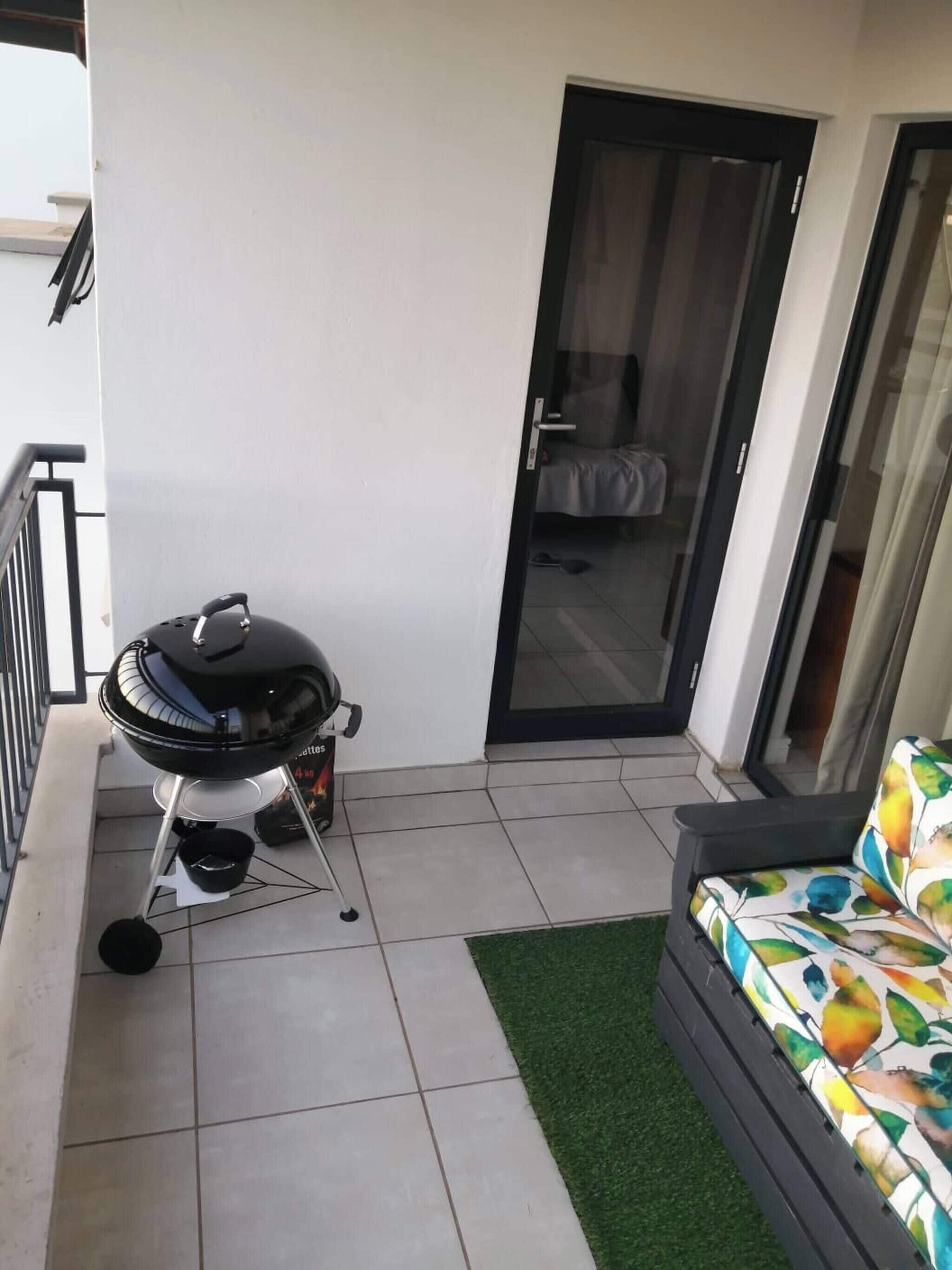 Un balcón con una parrilla, una puerta corredera de vidrio y un sofá con un cojín de colores.