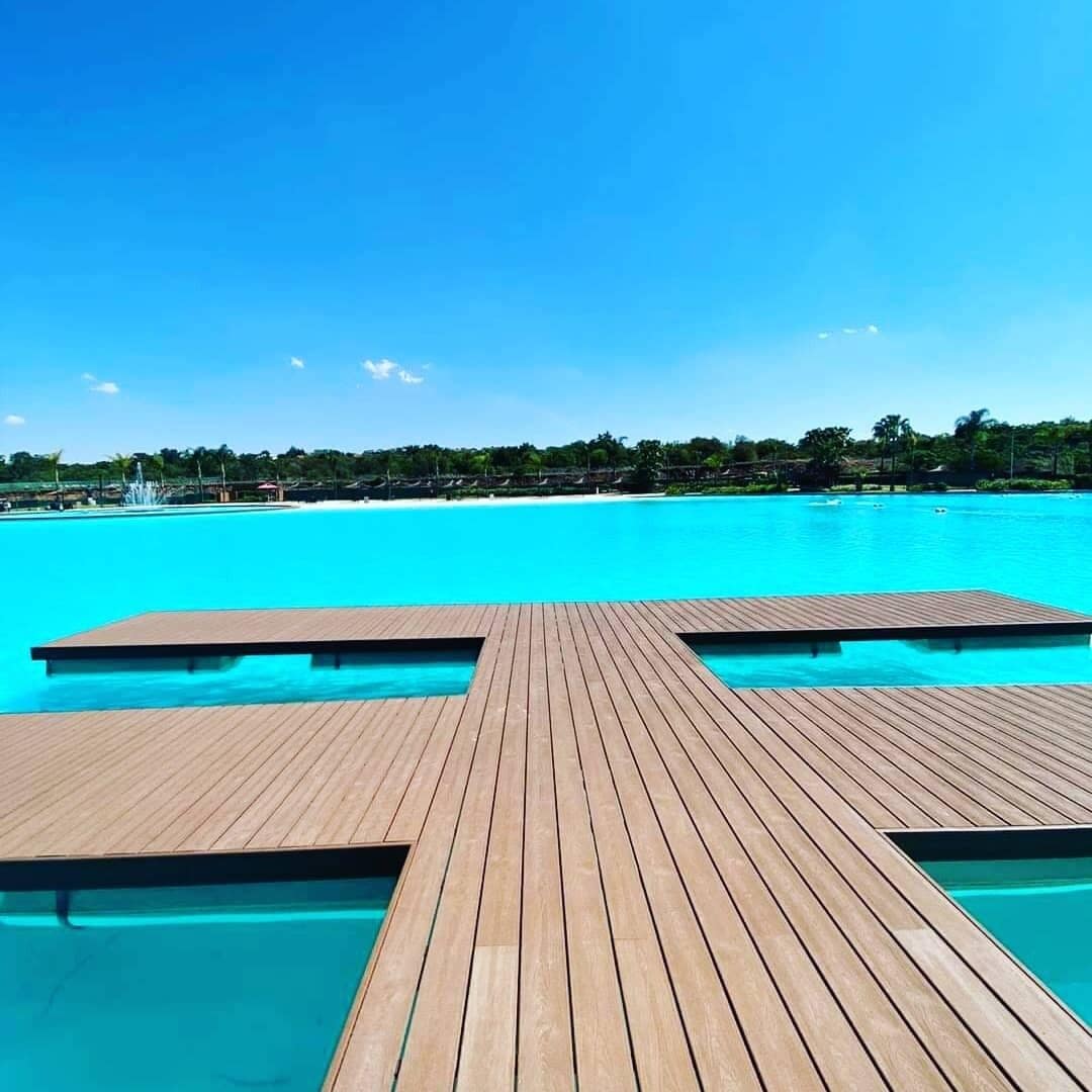 A wooden deck leading into a clear blue pool with a view of a beach and palm trees in the background.