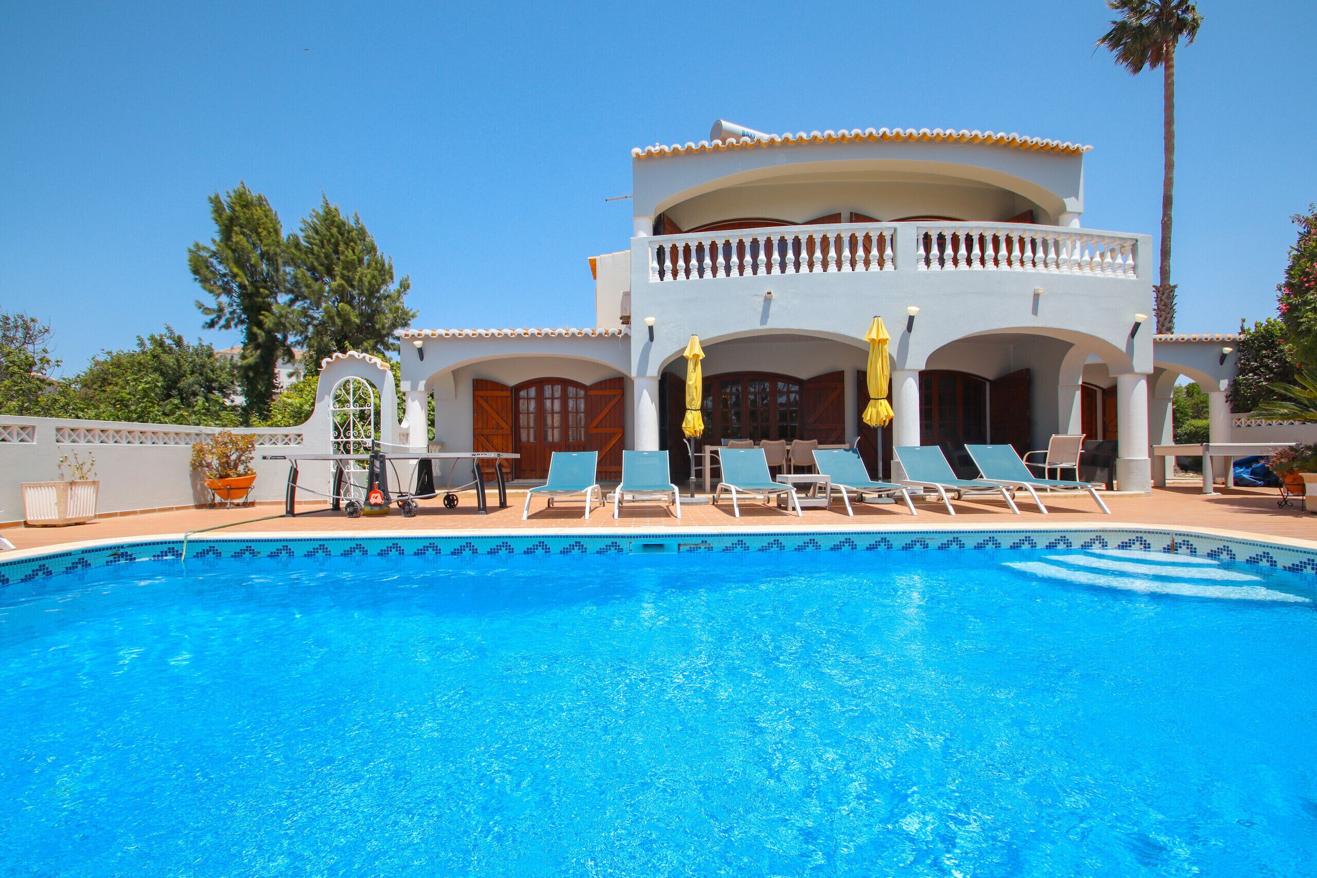 A pool with lounge chairs and umbrellas, a two-story building with balconies, and a clear blue sky.