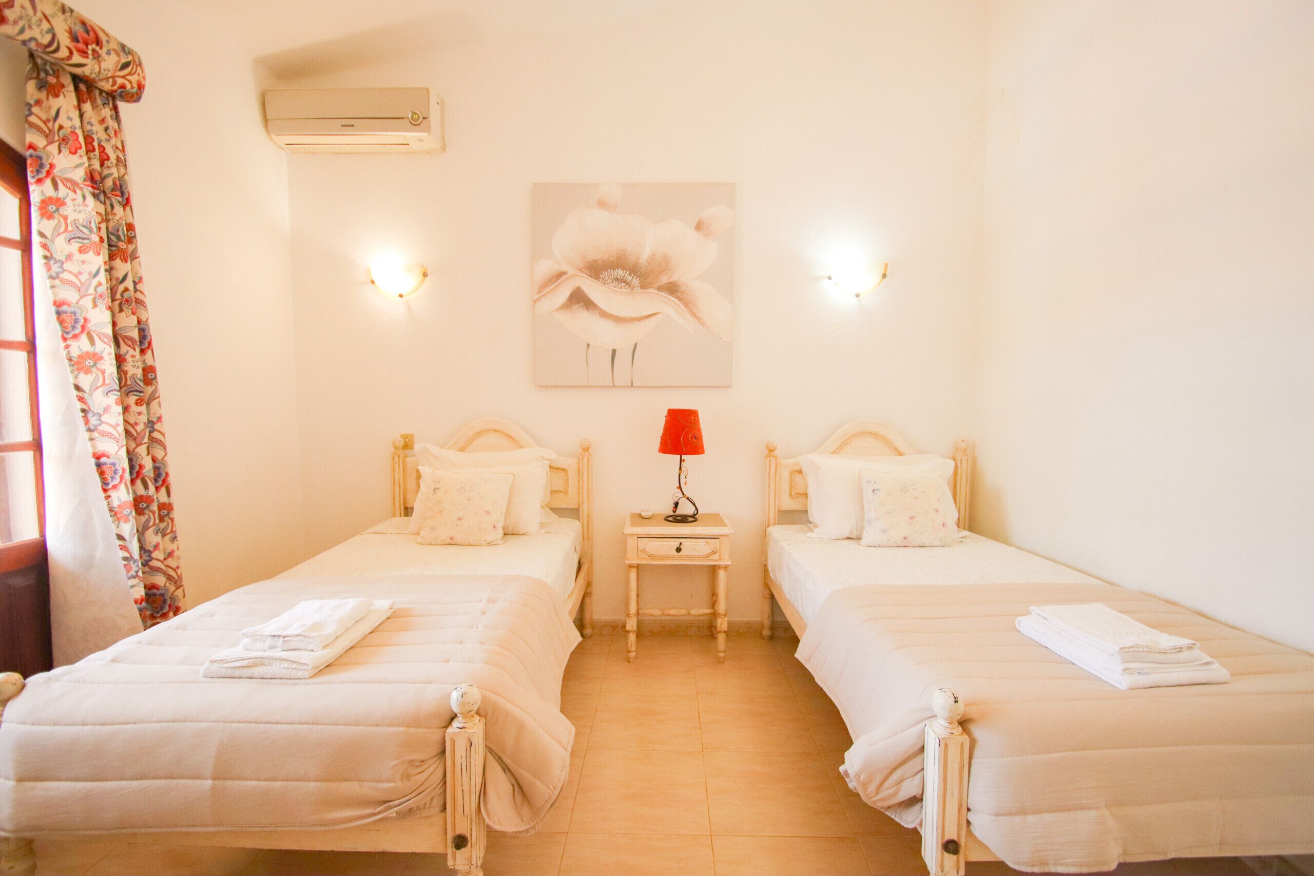 A room with two single beds, a nightstand with a lamp, an air conditioner, and a wall-mounted light fixture.
