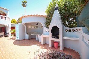 A courtyard with a barbecue area, a white building with a tiled roof, and a palm tree. - Villa with heatable pool and BBQ in Carvoeiro (Carvoeiro)