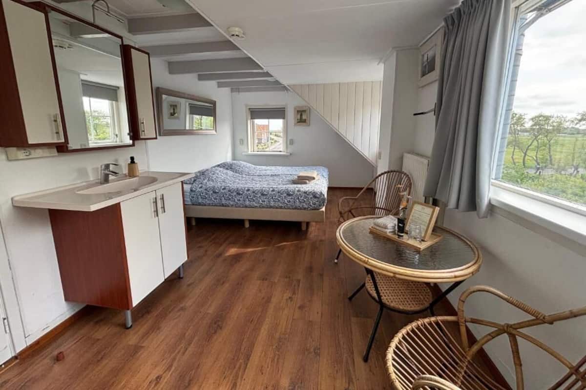 A bedroom with a bed, a small table with chairs, a window with curtains, a mirror, and a kitchenette with a sink and cabinets.