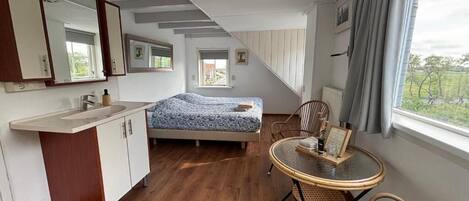 Un dormitorio con cama, una mesita con sillas, una ventana con cortinas, un espejo y una kitchenette con fregadero y armarios.