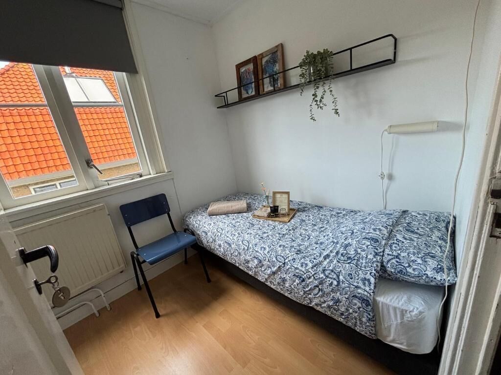 Een kleine, compacte kamer met een enkel bed, een stoel, een radiator, een raam met uitzicht op oranje daken en een plank met foto's en een plant.