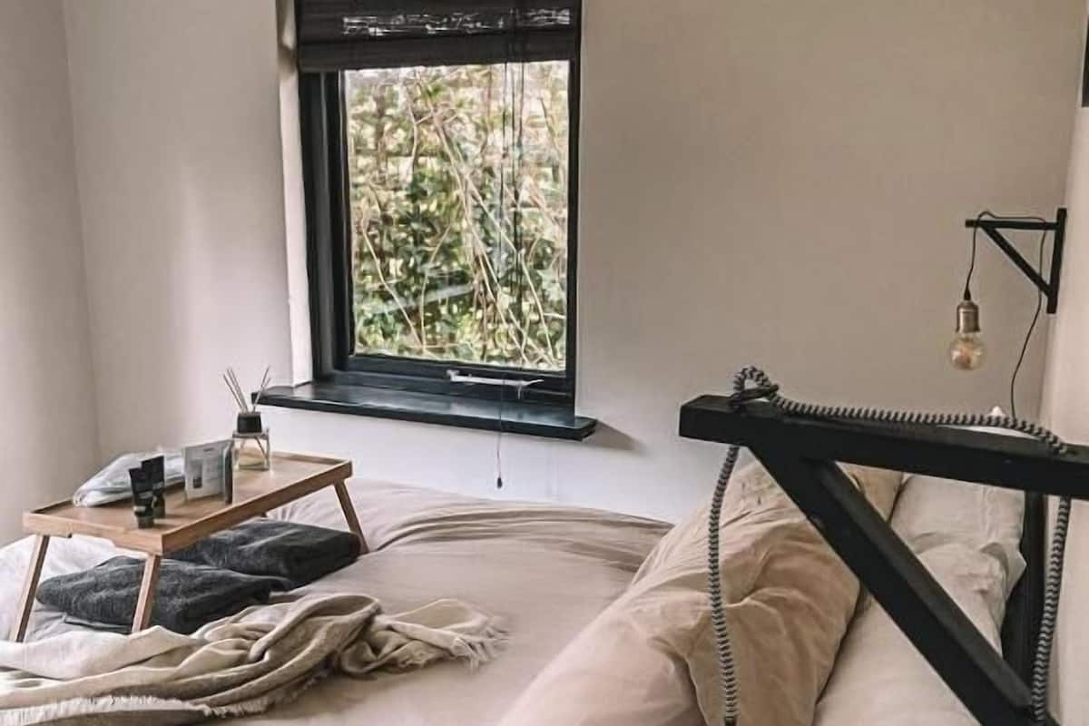 A bedroom with a bed, a window with a view of greenery, a bedside table with a lamp, and a hanging light fixture.