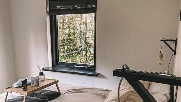 Quarto com cama, janela com vista para vegetação, mesa de cabeceira com luminária e luminária pendente.