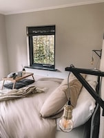 Un dormitorio con cama, una ventana que da a zonas verdes, una mesita de noche con lámpara, y una lámpara colgante.