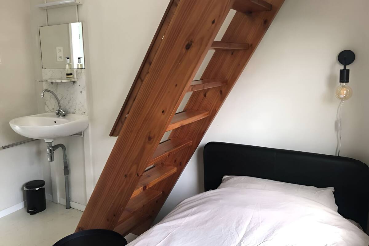 A bedroom with a black headboard, a wooden ladder leaning against the wall, a white sink, and a mirror above the sink.