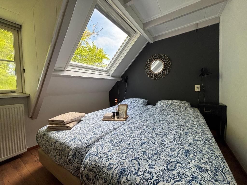 A bedroom with a skylight, a bed with a patterned bedspread, a bedside table, a mirror, and a lamp.