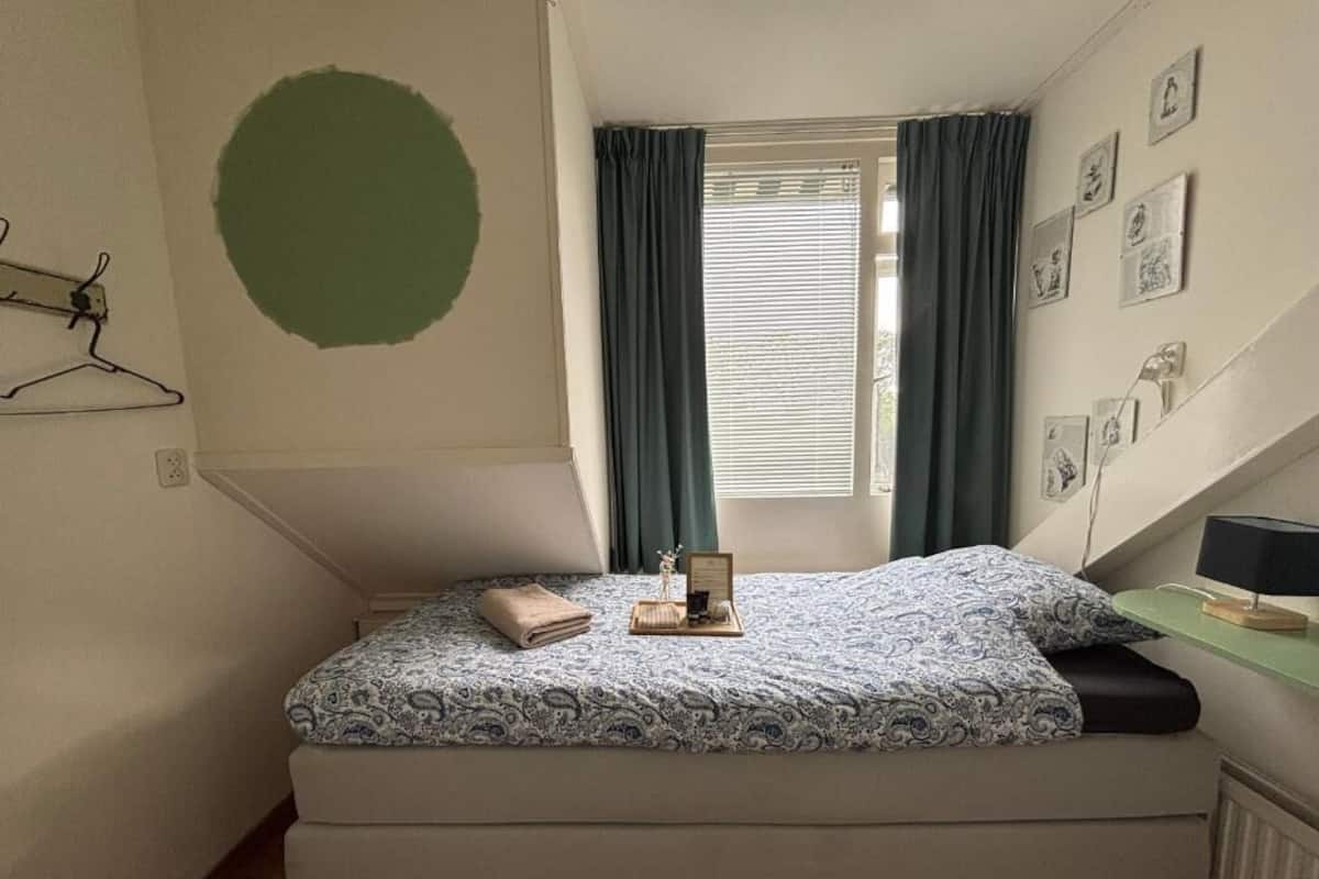 A small bedroom with a bed, a window with curtains, a wall-mounted light, and a radiator.