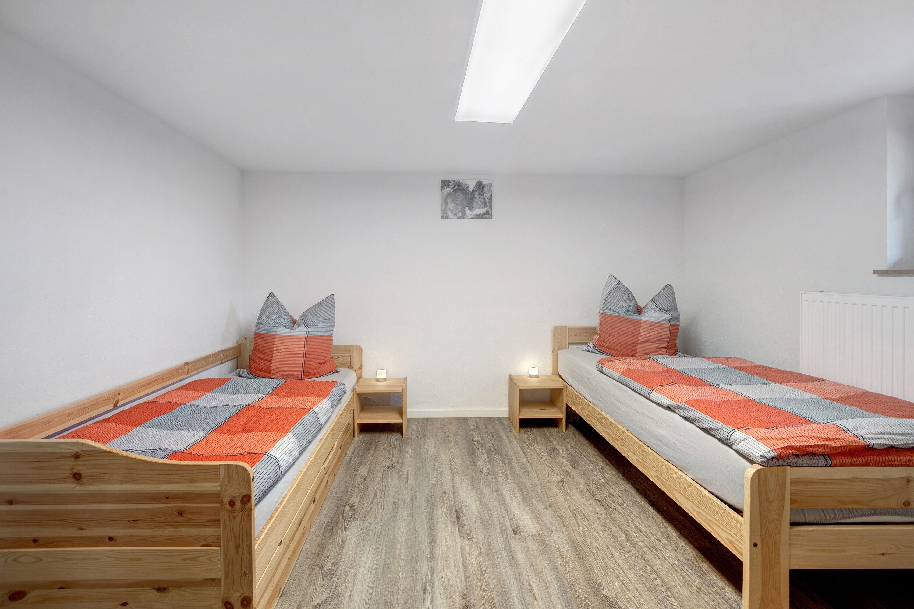 Ein Zimmer mit zwei Holzbetten, jeweils mit Kissen und Decke, einem kleinen Tisch, einem an der Wand hängenden Bild und einem Heizkörper.