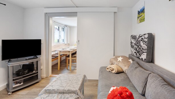 A modern living room with a flat-screen TV, grey sofa, and a dining area visible through an open door.
