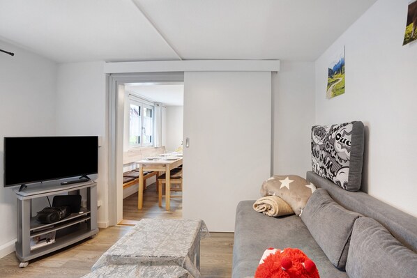 Un salon moderne avec un téléviseur à écran plat, un canapé gris et un coin repas visible à travers une porte ouverte.
