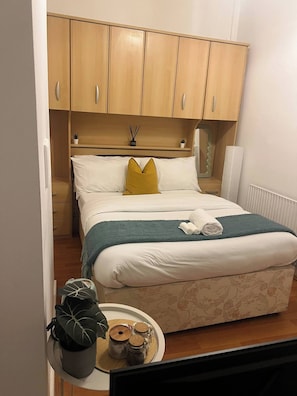 A bedroom with a bed, a small table with jars, a potted plant, and wooden cabinets above the bed. - Room-cozy Guest Room in Central Manchest (Manchester)