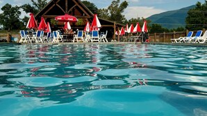 A swimming pool with lounge chairs and umbrellas, a wooden building, and a mountainous backdrop. - Luxurious Smoky Mountain Getaway Hot Tub 💦 Pool Sauna & Fishing 🐠 EV Charge ⚡️ (Sevierville)