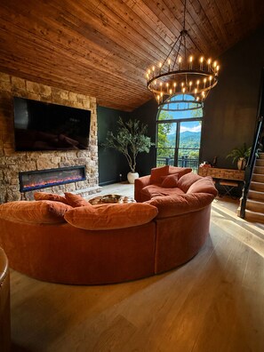 Ein modernes Wohnzimmer mit einer großen Eckcouch, einem Kamin, einem Fernseher und Blick ins Freie.