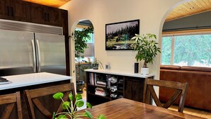 A kitchen with a wooden table, chairs, a refrigerator, and a dining area with a view of the outdoors. - Cozy Mountain Retreat | Steps to Bow River (Canmore)