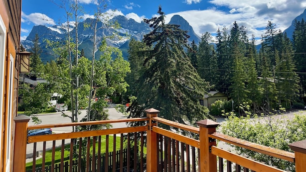 A wooden balcony with a view of a mountain range, a road, and a green landscape. - Cozy Mountain Retreat | Steps to Bow River (Canmore)