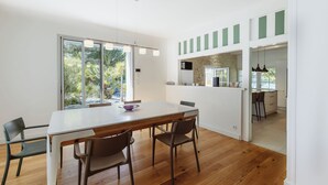 Een moderne keuken met een groot eiland, een eethoek met een witte tafel en stoelen, en een uitzicht op de omgeving door grote ramen.