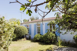 Una casa con fachada blanca y contraventanas azules, rodeada de vegetación y un cielo despejado.