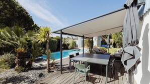 Een terras buiten met een zwembad, een overdekte zithoek en een tafel.