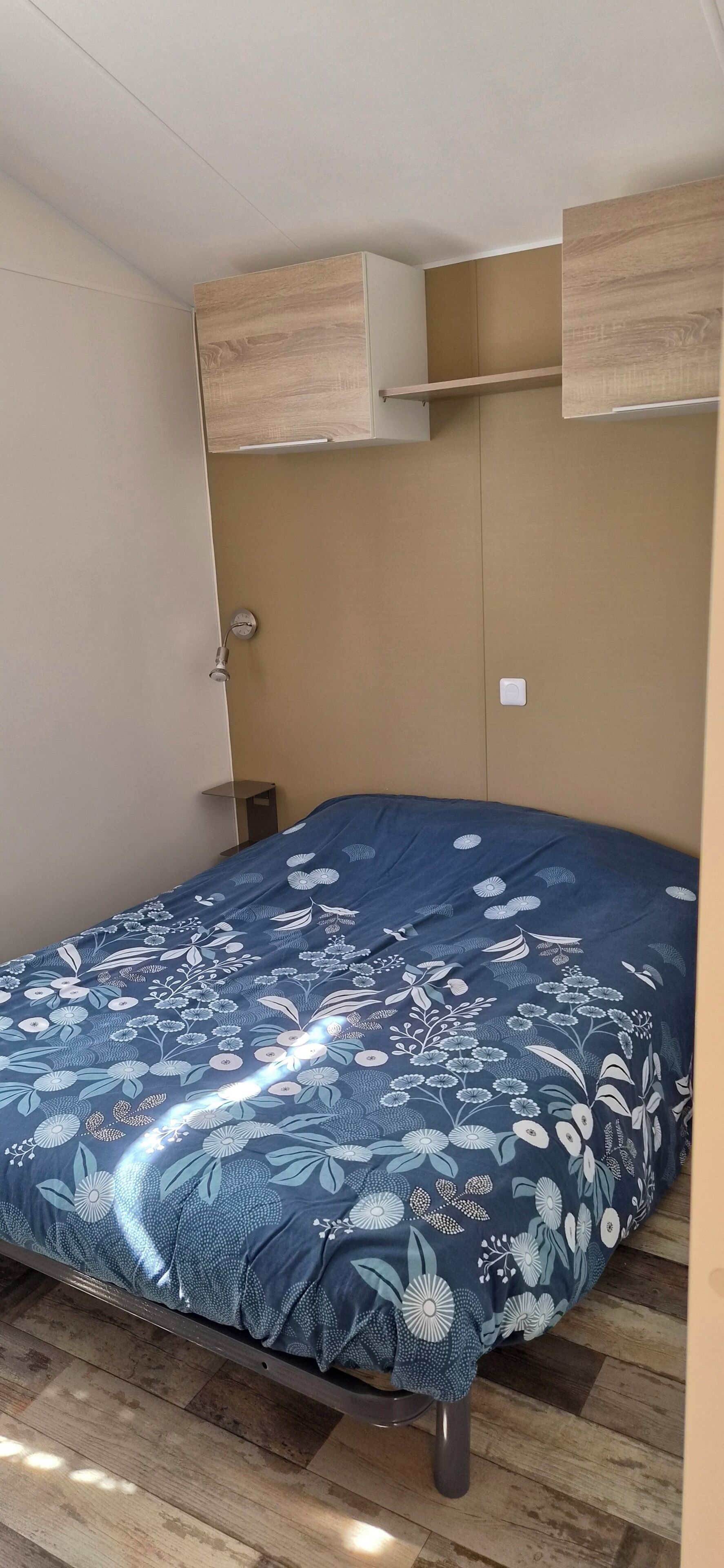 Ein Zimmer mit einem Bett, einem hölzernen Kopfende, einem Nachttisch und einem an der Wand befestigten Regal.