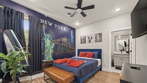 A modern bedroom with a cityscape mural, a bed with blue bedding and red pillows, a bench, and a ceiling fan. - Sleep In Style Fashion House W/Pool Table Sleep 10 (Houston)
