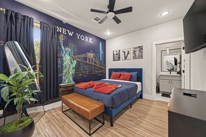 Ein modernes Schlafzimmer mit Stadtbild-Wandgemälde, einem Bett mit blauer Bettwäsche und roten Kissen, einer Bank und einem Deckenventilator.