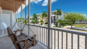 A wooden swing chair on a covered balcony with a view of a residential area, palm trees, and a clear blue sky. - 🌊 Palermo Paradise | 3BR Fort Myers Beach | Heated Pool + Porch Swing (Fort Myers Beach)
