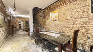 Un comedor con pared de ladrillo, mesa de vidrio, sillas de metal y una escalera que sube a otra habitación.