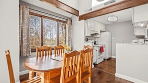 Ein Essbereich mit Holztisch und -stühlen, einem Fenster mit Vorhängen und einer Küche mit weißen Schränken und Geräten.