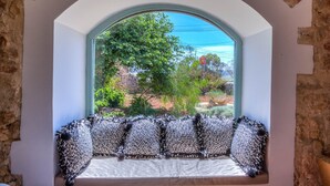 Una habitación con pared de piedra, ventana con vistas a la vegetación y un banco con cojines estampados.