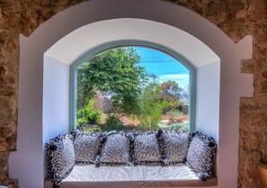 A room with a stone wall, a window with a view of greenery, and a bench with patterned cushions. - Wonderful 4BR Farmhouse with Private Pool (Rabat)