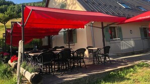 An outdoor seating area with tables and chairs under red umbrellas, a building with a sloped roof, and a wooden deck. - Auberge du Graber (Dolleren)