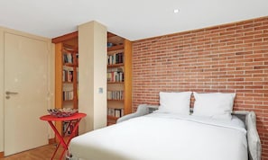 Ein Schlafzimmer mit einem Bett, einem roten Tisch und Bücherregalen an einer Ziegelwand.