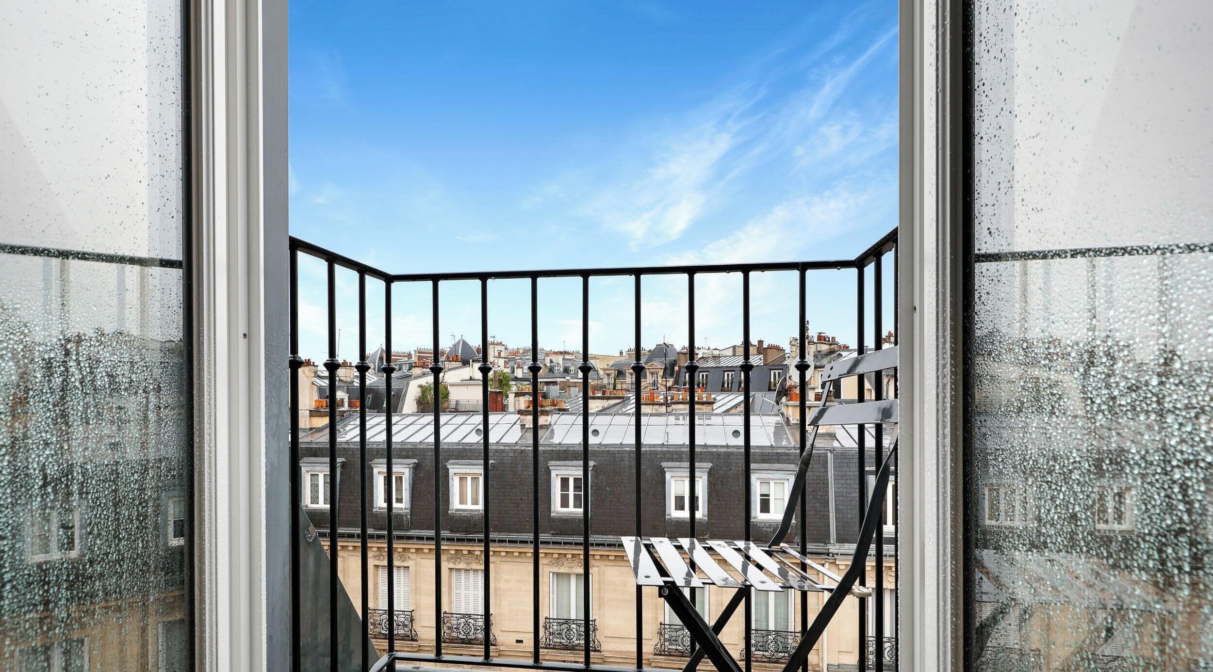 A view through a barred window with a clear sky, revealing a cityscape with multiple buildings and rooftops.