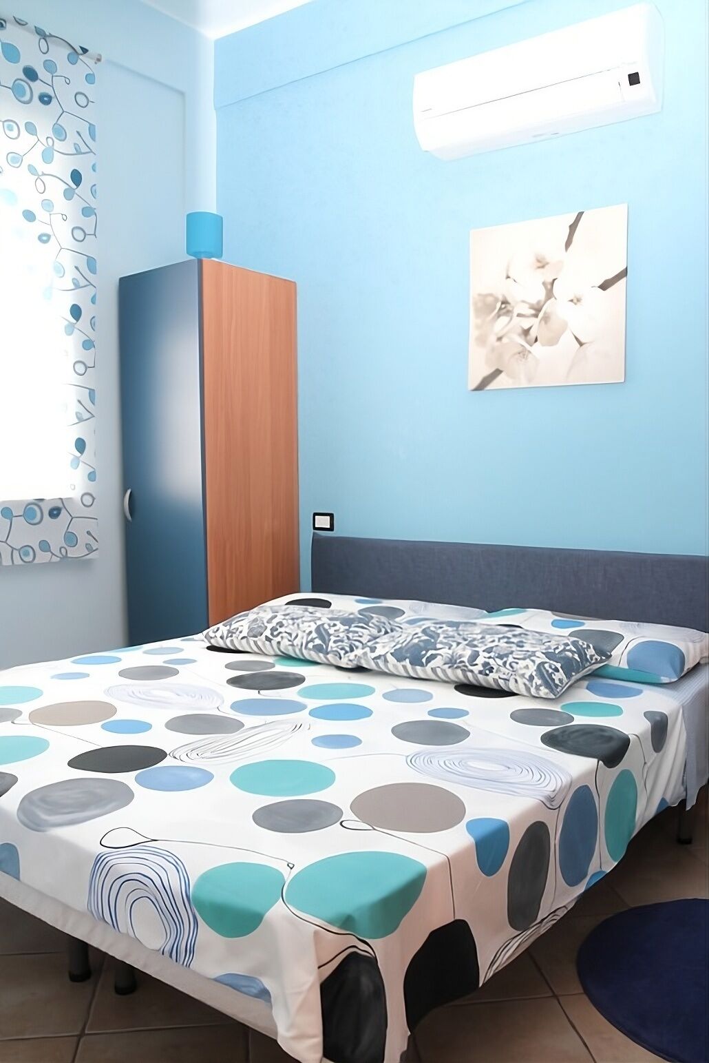 A bedroom with a bed, a wardrobe, a wall-mounted air conditioner, and a window with a curtain.