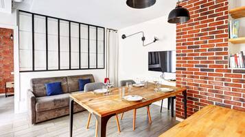Een moderne woonkamer met een houten eettafel voor zes personen, een bank, een bakstenen muur en een groot raam.