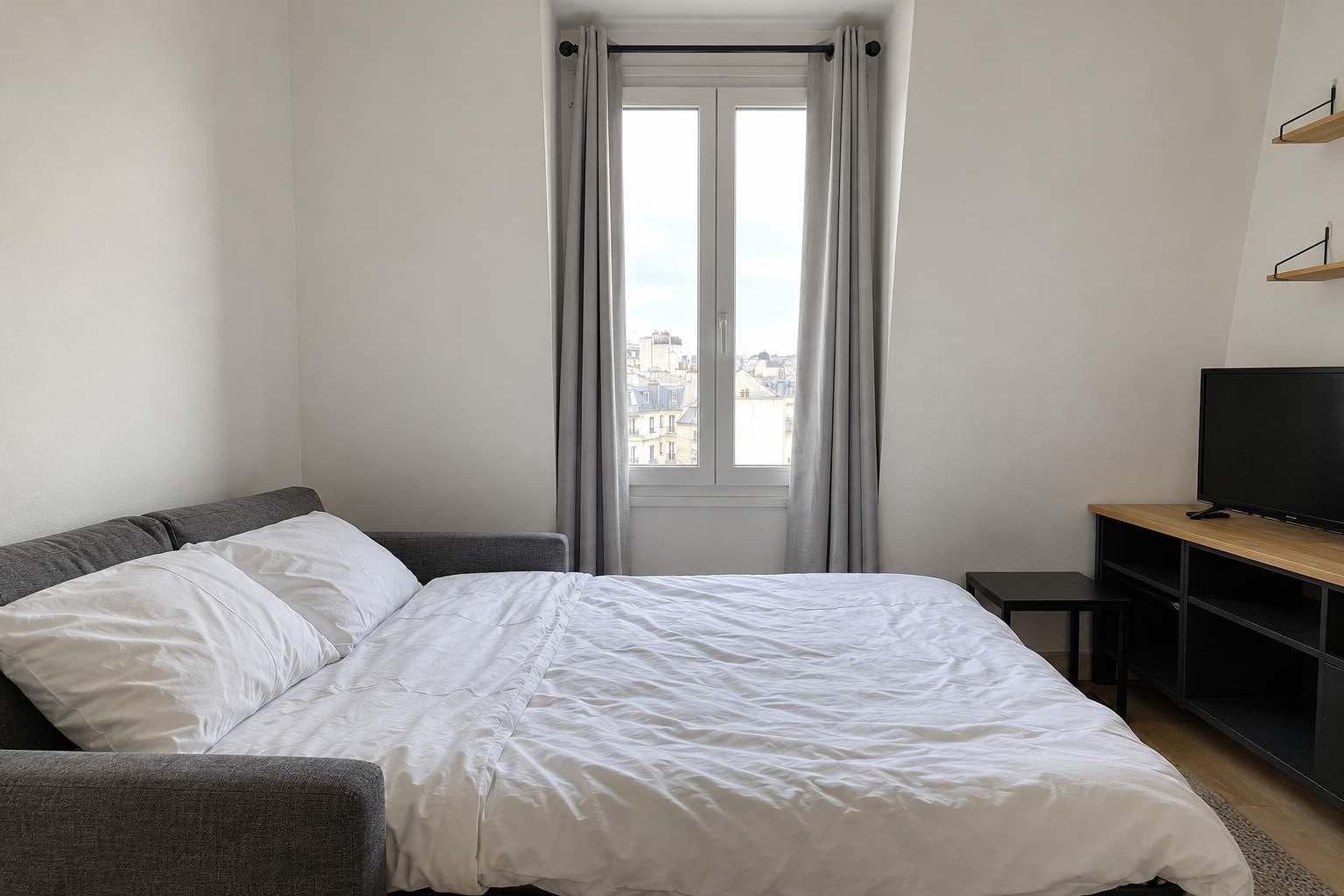 Una habitación de hotel con una cama, un televisor sobre un pedestal de madera y una ventana con cortinas.