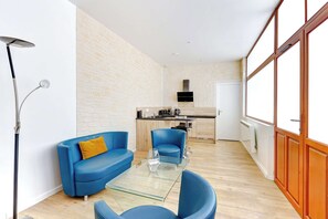 Ein modernes Wohnzimmer mit einem Glastisch, blauen Sofas und einer Küche im Hintergrund.