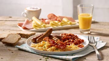 Ein Frühstücksteller mit Eiern, Würstchen, Bohnen und Toast, ein Glas Orangensaft und eine Tasse Kaffee auf einem Holztisch.